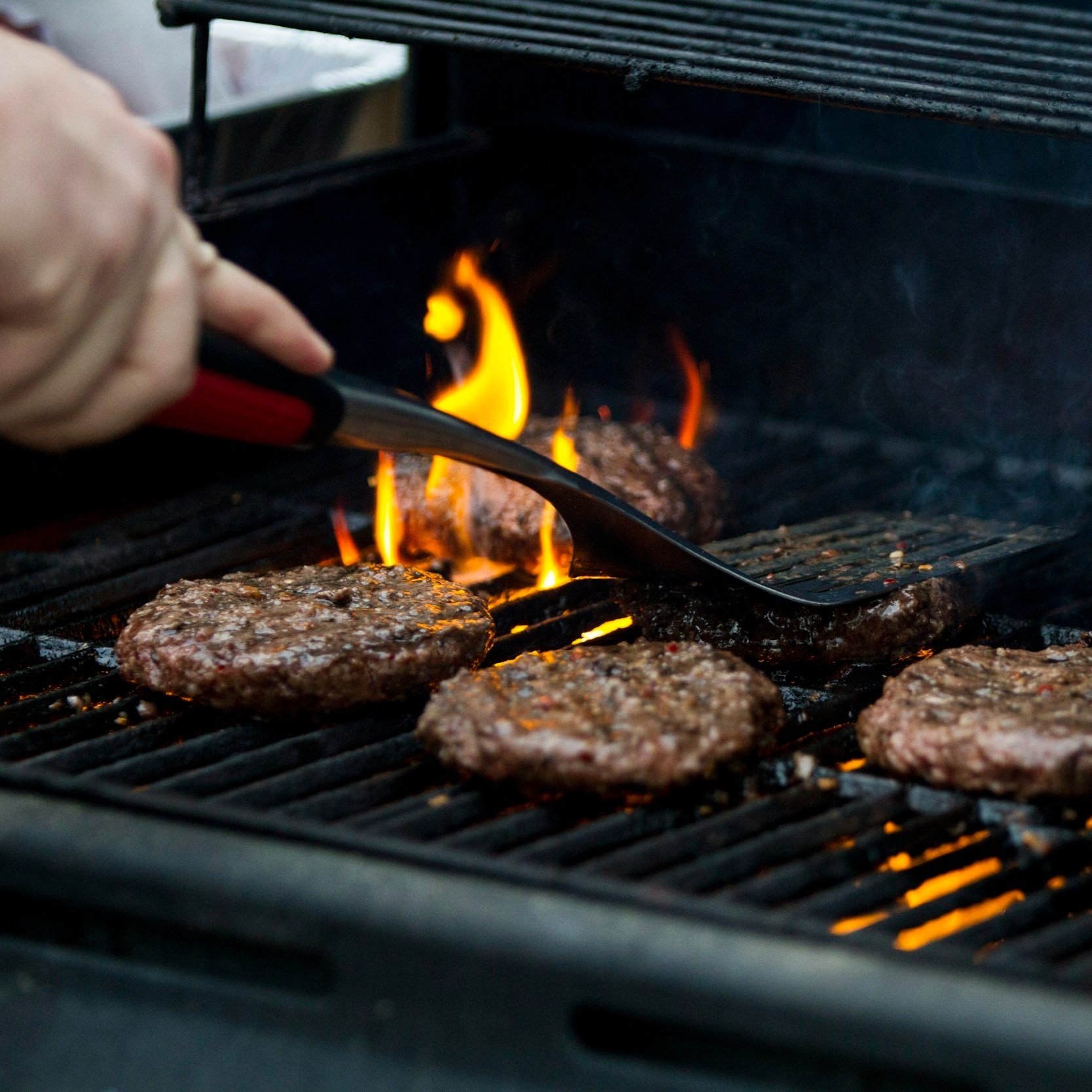 Close-up of a hand using a spatula to flip sizzling beef burgers on a grill, with flames rising from the heat, showcasing outdoor cooking and barbecue preparation.